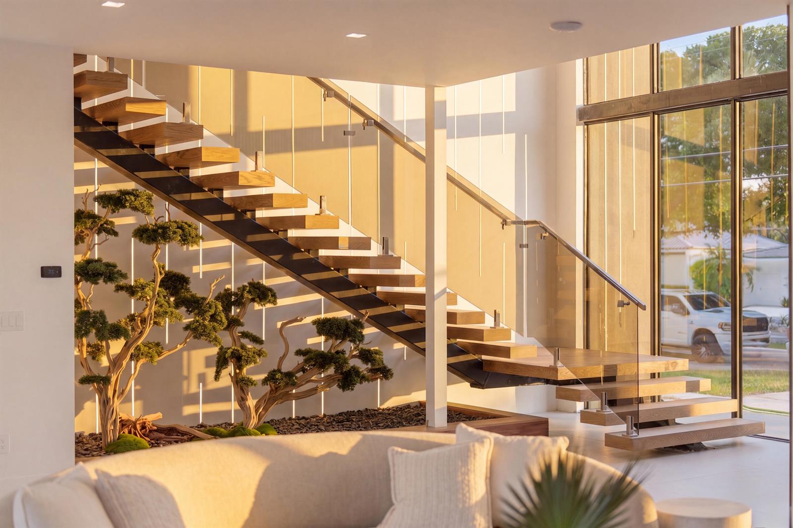 A modern living room features a floating wooden staircase and indoor bonsai trees near windows — 2360 NE 49th Street, Lighthouse Point FL 33064, LIGHTHOUSE POINT, FL