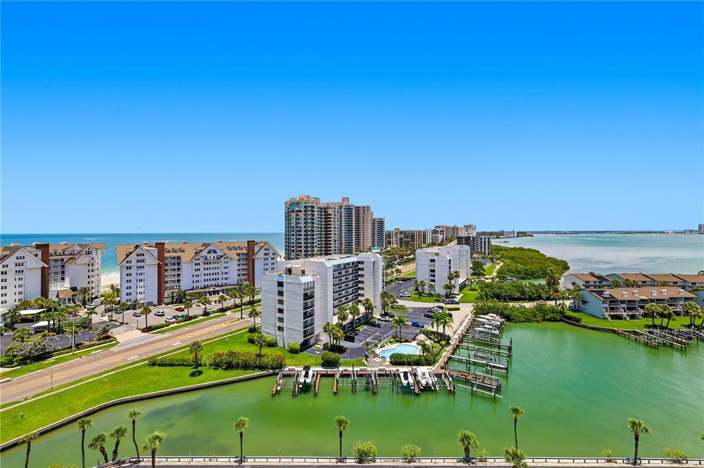 An aerial view shows waterfront condominiums, a boat dock, and a road near the ocean — 1621 GULF BOULEVARD UNIT 1408, CLEARWATER BEACH, FL