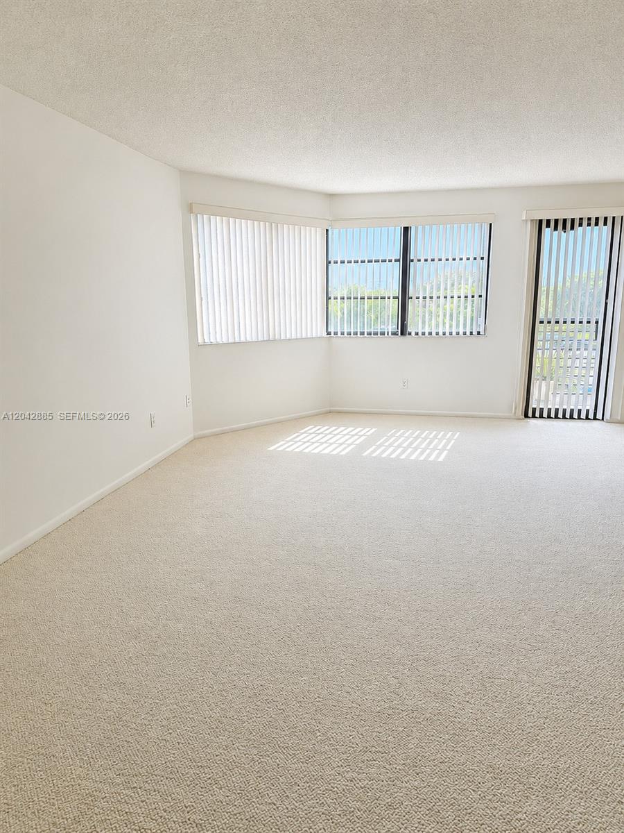 An empty living room features beige carpeting, white walls, and large windows with vertical blinds — 20840 San Simeon Way # 308, Miami FL 33179, MIAMI, FL