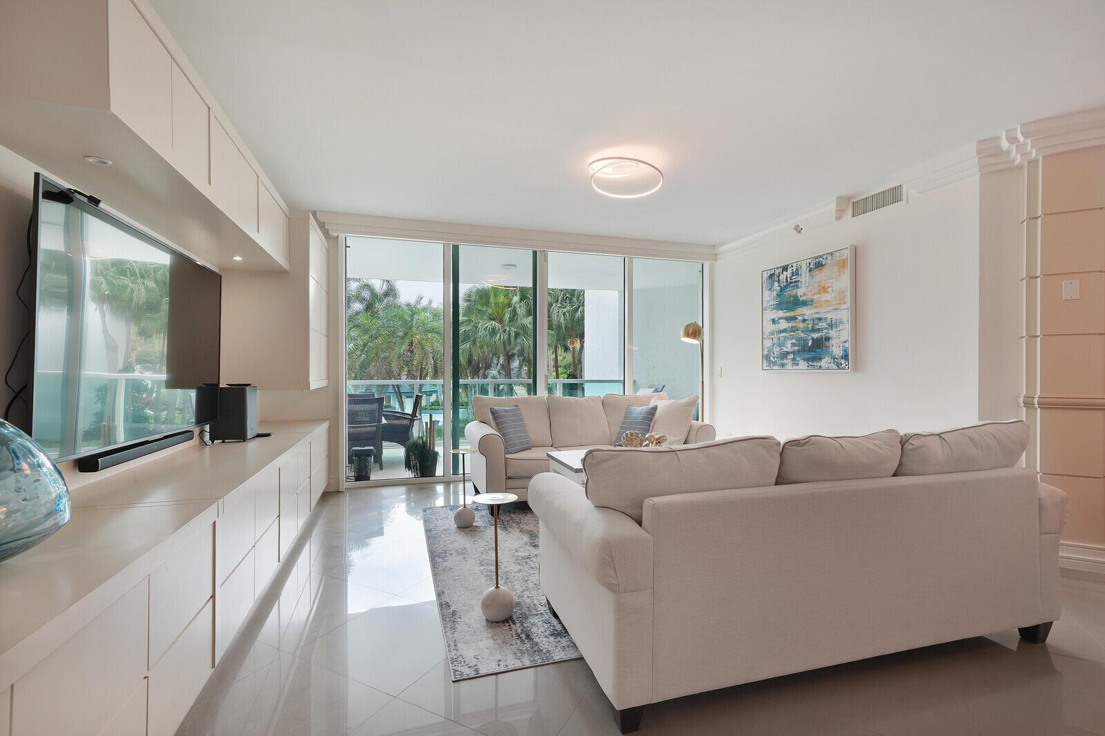 A modern living room features a large white sofa, built-in cabinetry, and a glass balcony door — 550 SE Mizner Boulevard # B209, Boca Raton FL 33432, BOCA RATON, FL