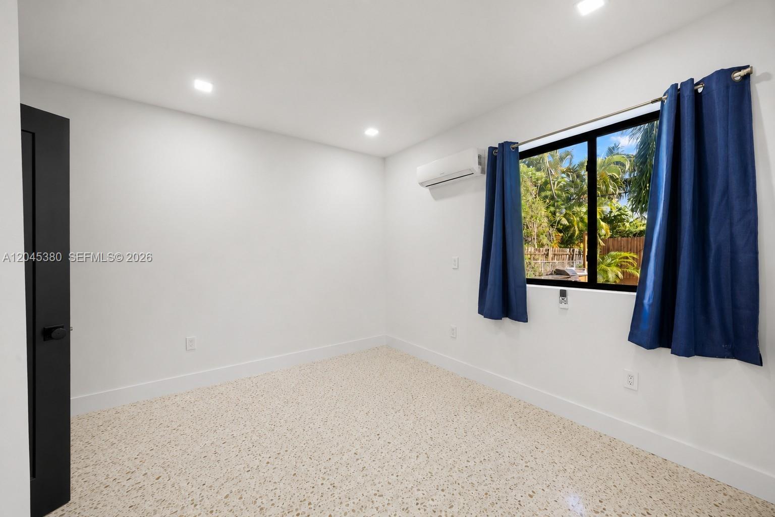 A bedroom features white walls, terrazzo flooring, a window with blue curtains, and a wall-mounted AC unit — 365 Fairway Dr, Miami Beach FL 33141, MIAMI BEACH, FL