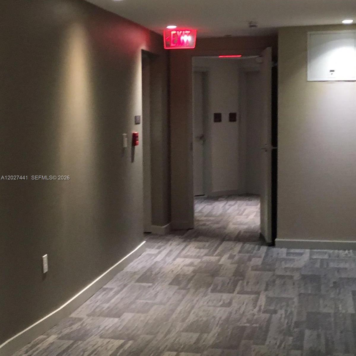 A hallway with gray patterned carpet leads to an open doorway with an exit sign — MIAMI, FL