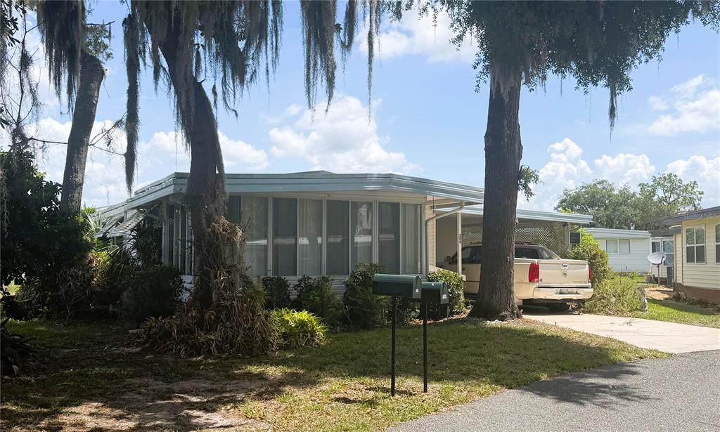 A single-story mobile home with a screened-in porch and a carport sits under large trees — 209 JACARANDA DRIVE, LEESBURG, FL