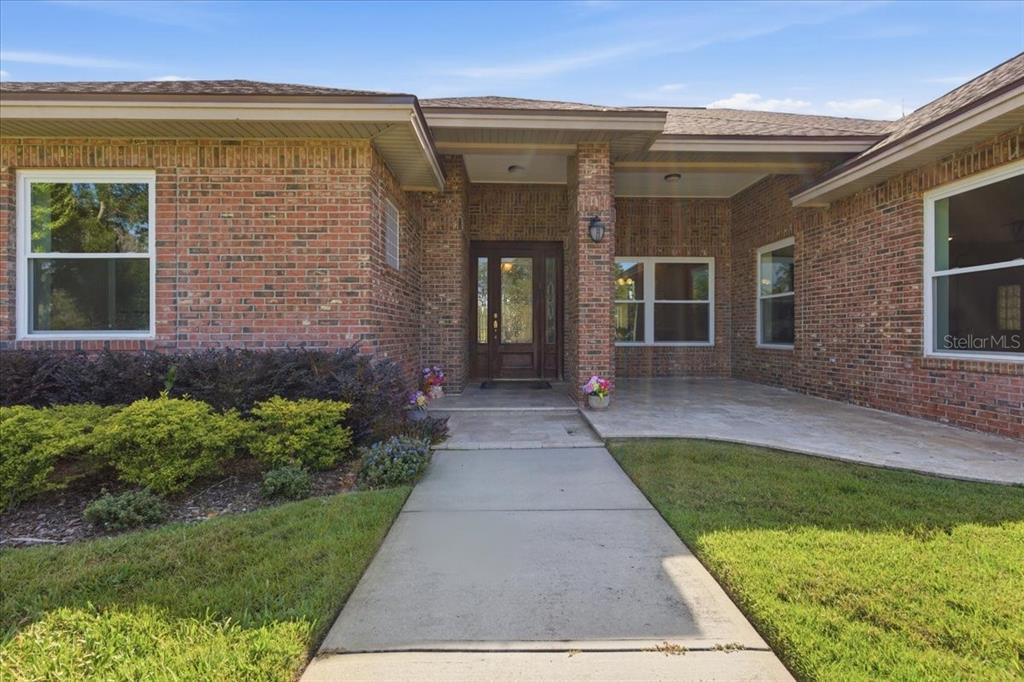 A brick house exterior features a covered entryway, a concrete walkway, and a landscaped lawn — 17825 SIMMS ROAD, ODESSA, FL