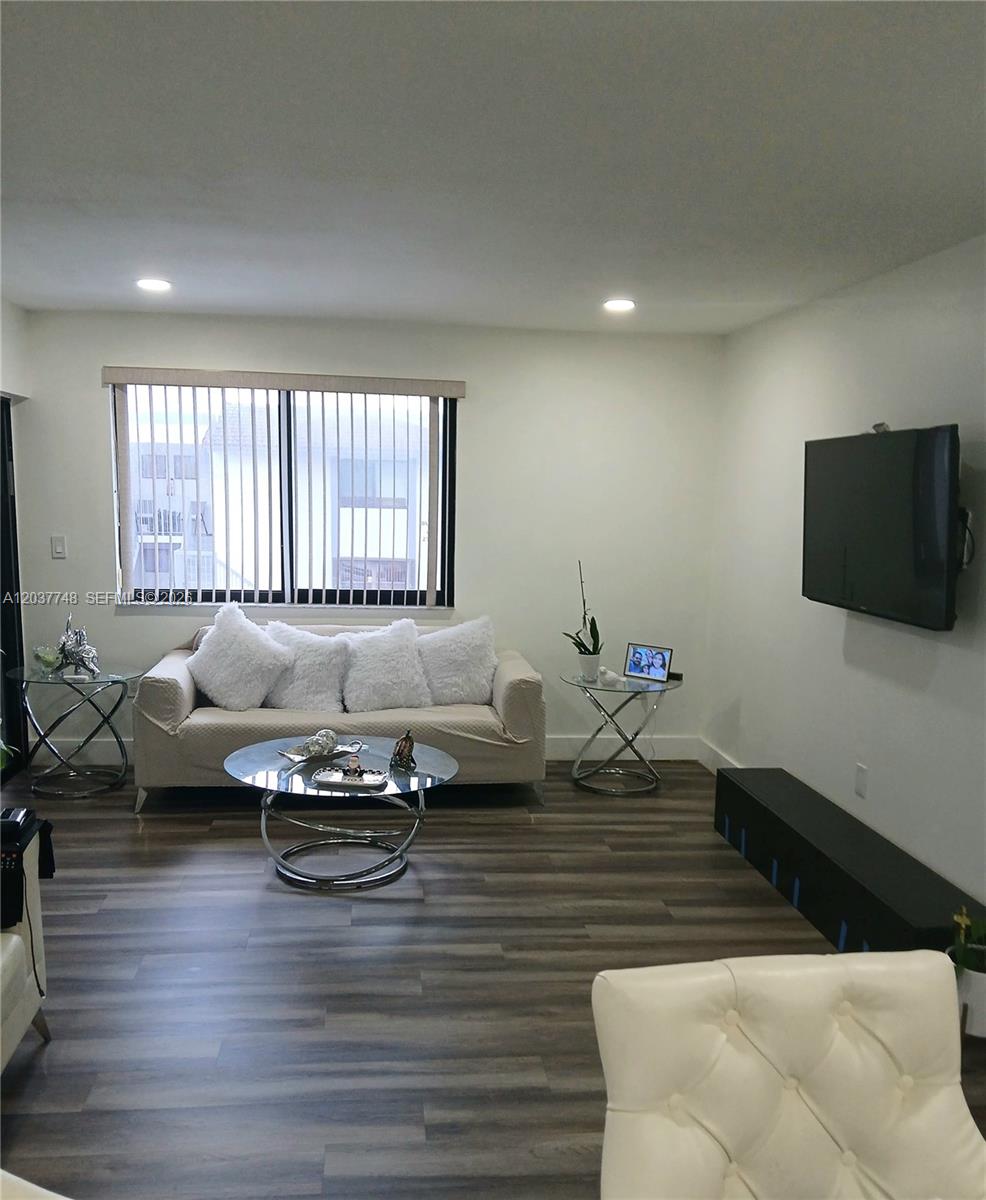 A living room features a beige sofa, glass tables, a wall-mounted television, and wood-look flooring — 6365 W 27th Ct # 8, Hialeah FL 33016, HIALEAH, FL