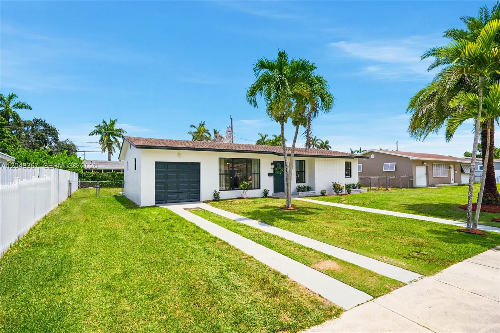 A white single-story house features a dark garage door, a front lawn, and palm trees — 14785 SW 297th St, Homestead FL 33033, HOMESTEAD, FL