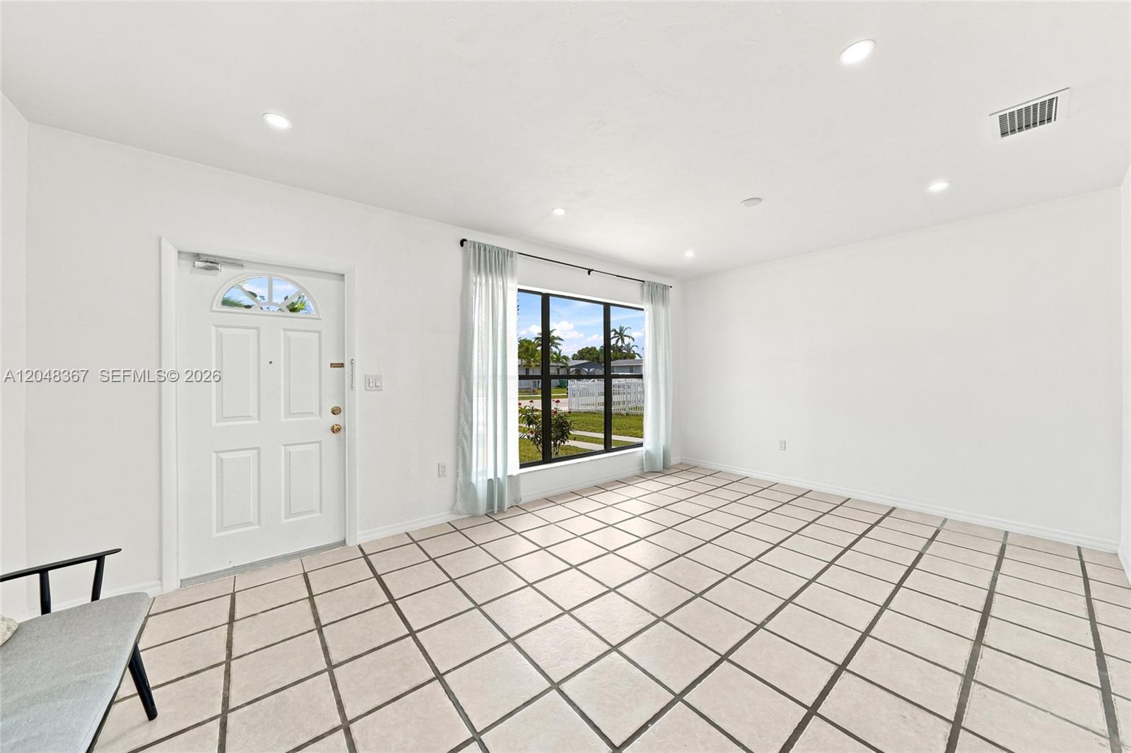 A bright, empty room features white walls, tiled flooring, a front door, and a large window — 14785 SW 297th St, Homestead FL 33033, HOMESTEAD, FL