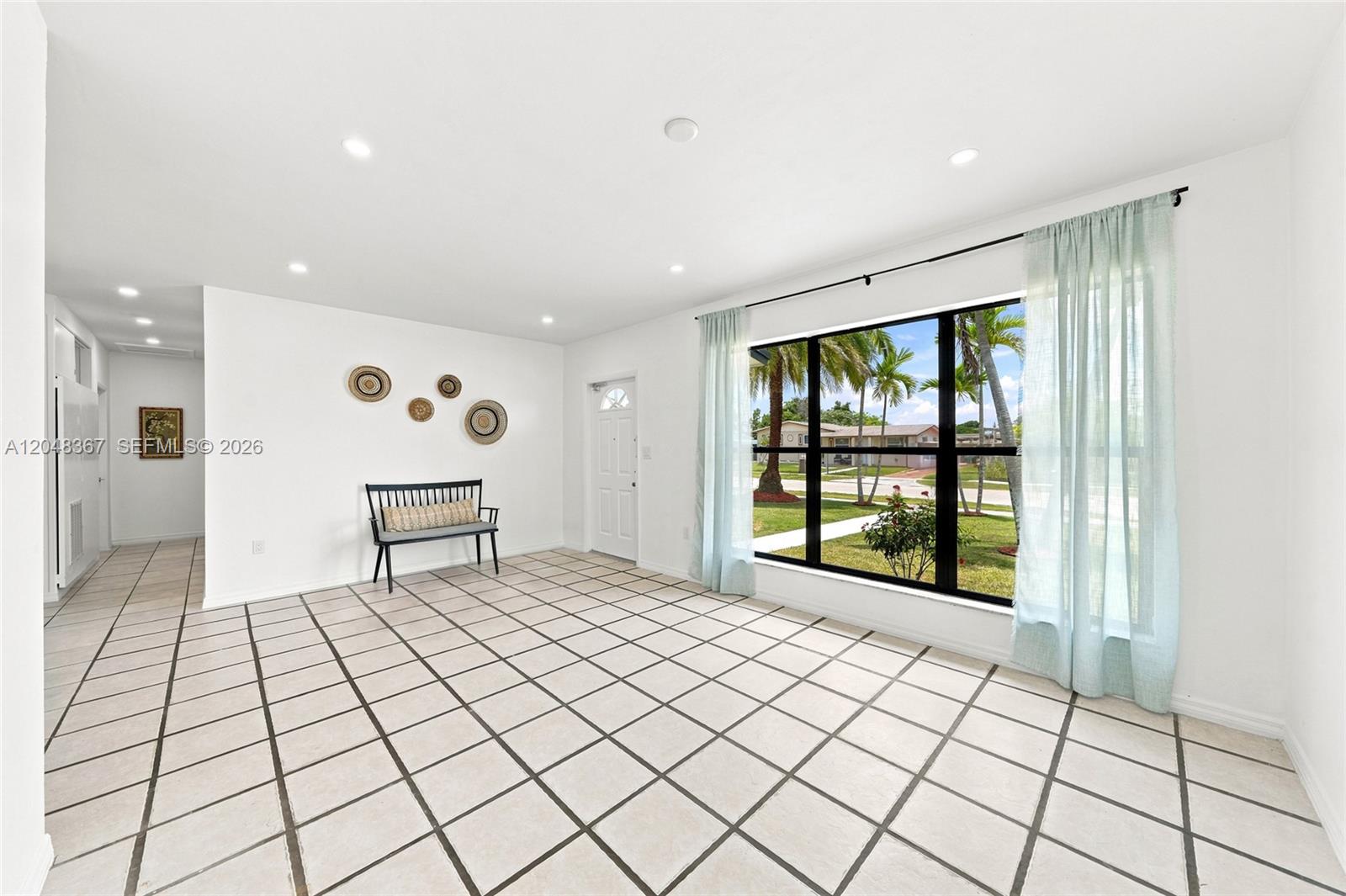 A bright, tiled living room features a black bench, wall decor, and a large window — 14785 SW 297th St, Homestead FL 33033, HOMESTEAD, FL