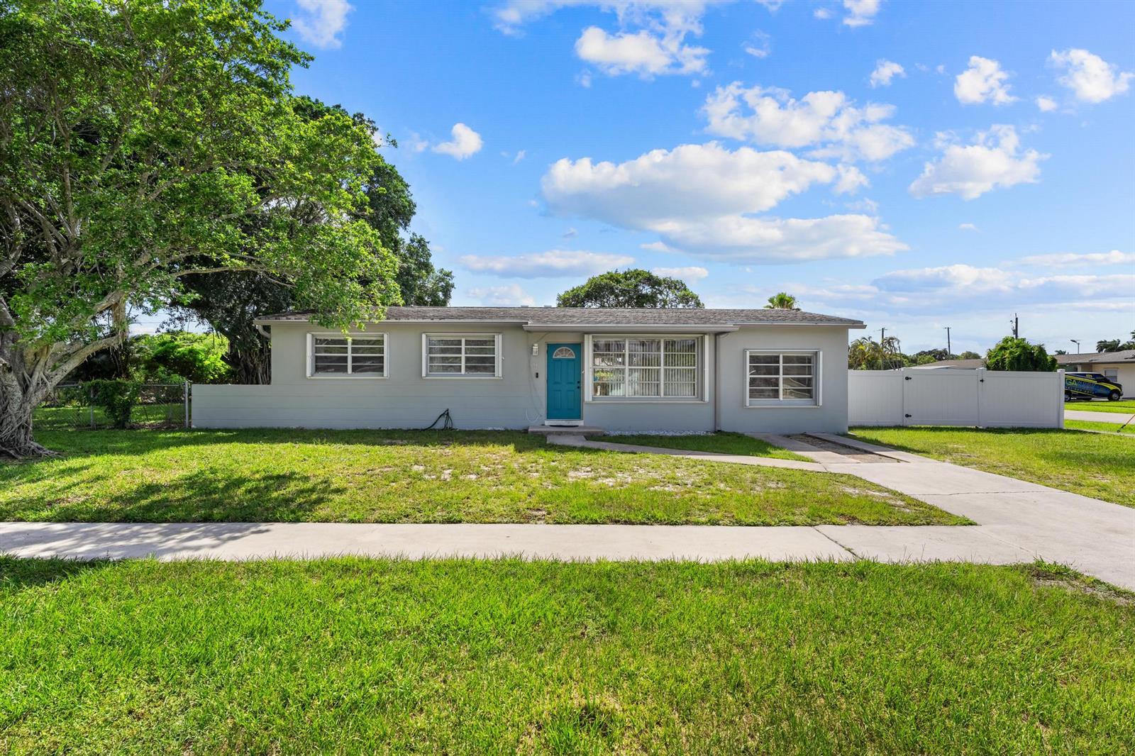 A single-story gray house with a blue front door sits behind a green lawn — 153 Abaco Drive, Palm Springs FL 33461, PALM SPRINGS, FL