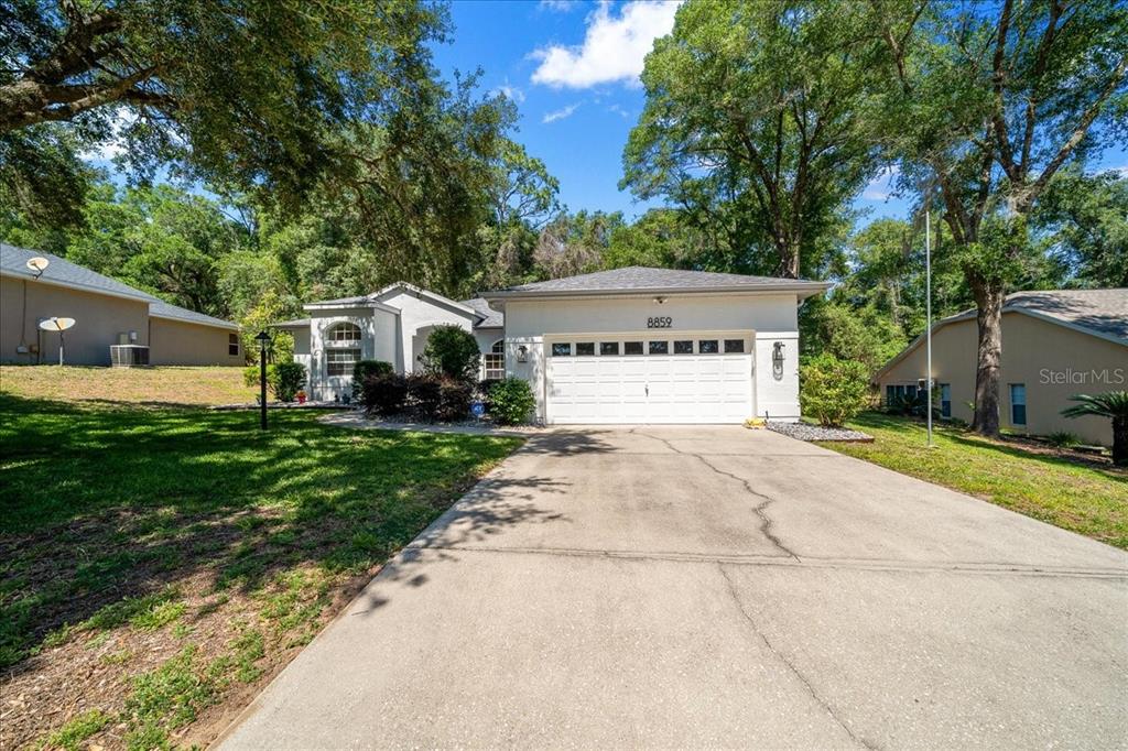 A single-story house with a white garage, concrete driveway, and surrounding trees under a blue sky — 8859 SW 196TH AVENUE ROAD, DUNNELLON, FL