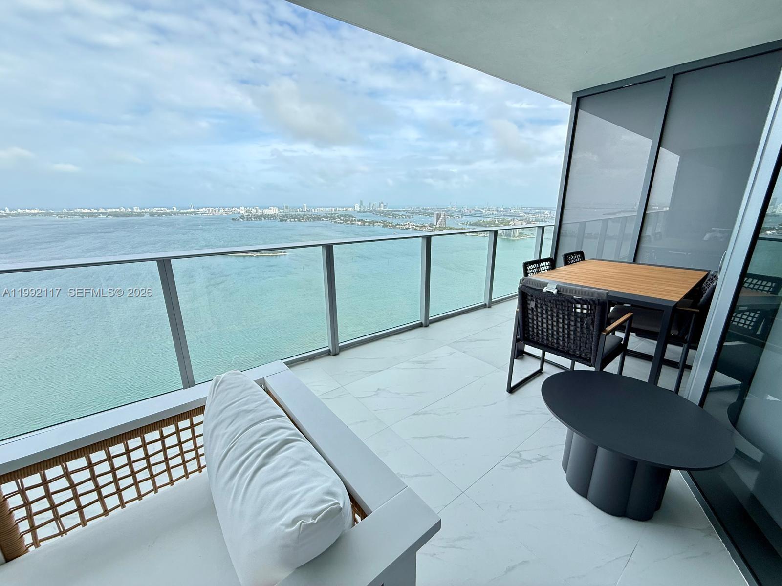 A high-rise balcony features white tile flooring, outdoor furniture, and a panoramic bay view — 700 NE 24 street # 3802, Miami FL 33137, MIAMI, FL