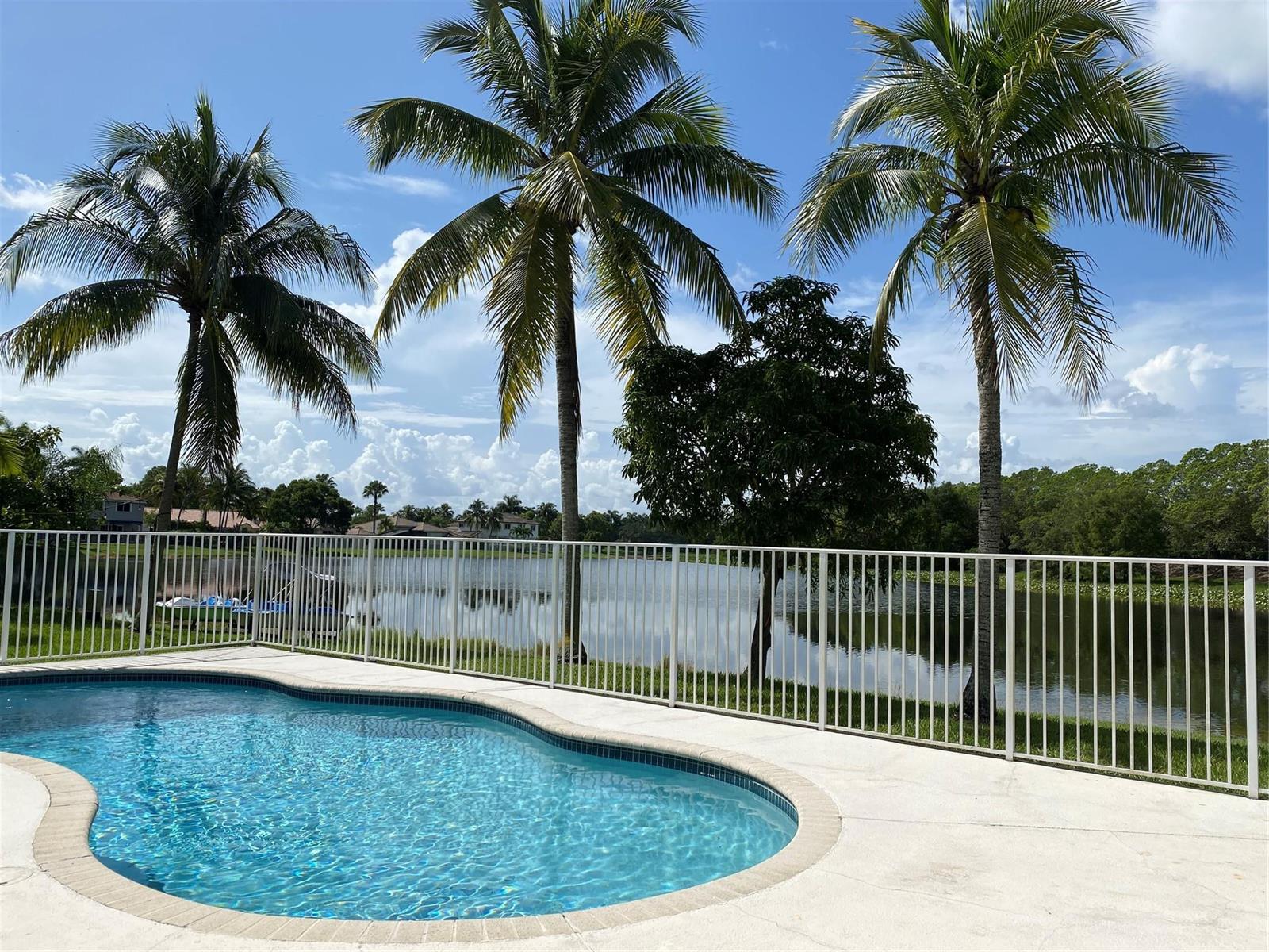 A swimming pool sits on a patio behind a white fence overlooking a lake — 338 Cambridge Drive, Weston FL 33326, WESTON, FL