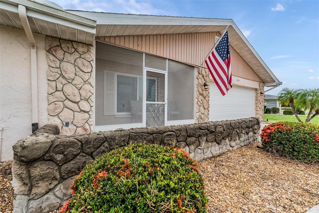 The exterior of a house features a stone-faced wall, a screened porch, and an American flag — 1209 WILD FEATHER LANE, SUN CITY CENTER, FL