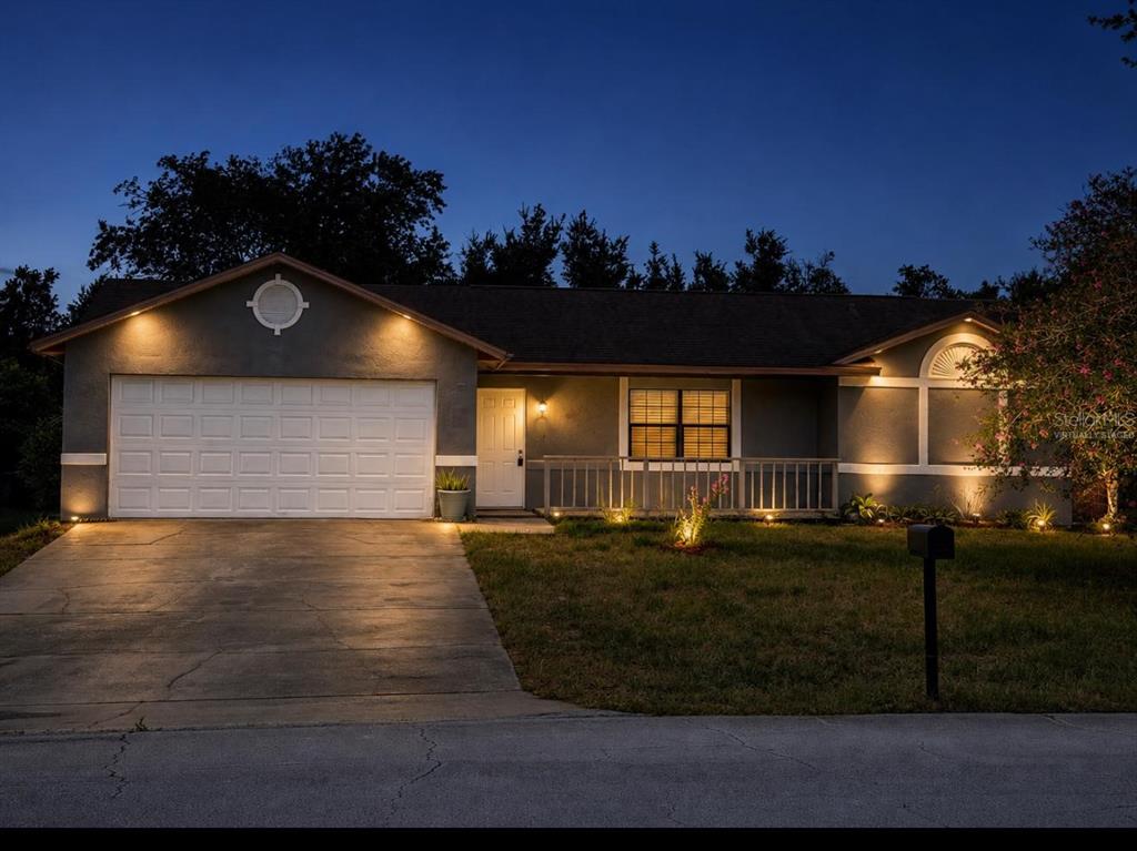 A single-story house at dusk with a garage, driveway, and illuminated exterior lighting — 1726 MONTICELLO STREET, DELTONA, FL