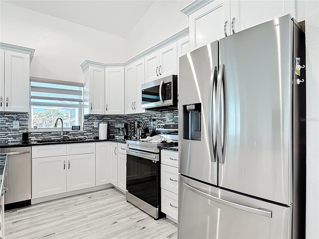 A kitchen features white cabinetry, stainless steel appliances, a black tiled backsplash, and wood-look flooring — 202 LONG MEADOW LANE, ROTONDA WEST, FL