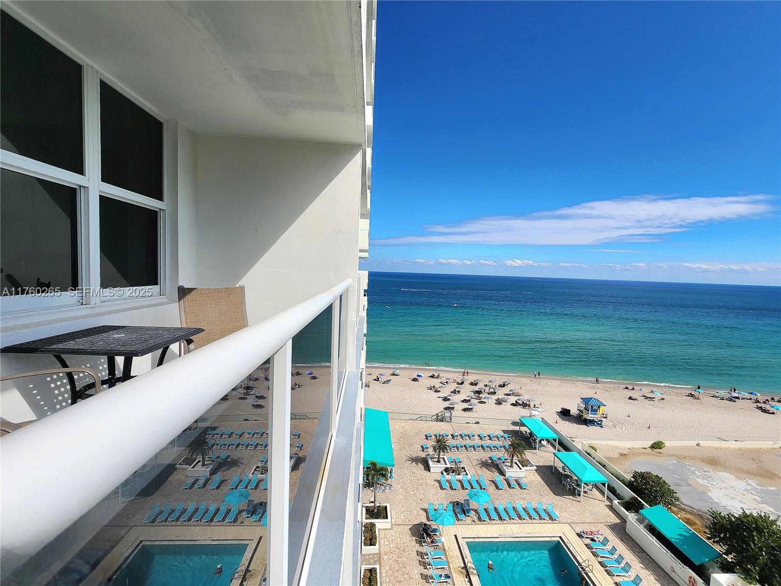 A high-rise balcony overlooks a swimming pool, beach, and the ocean under a clear sky — 3725 S Ocean Dr # 1008, Hollywood FL 33019, HOLLYWOOD, FL