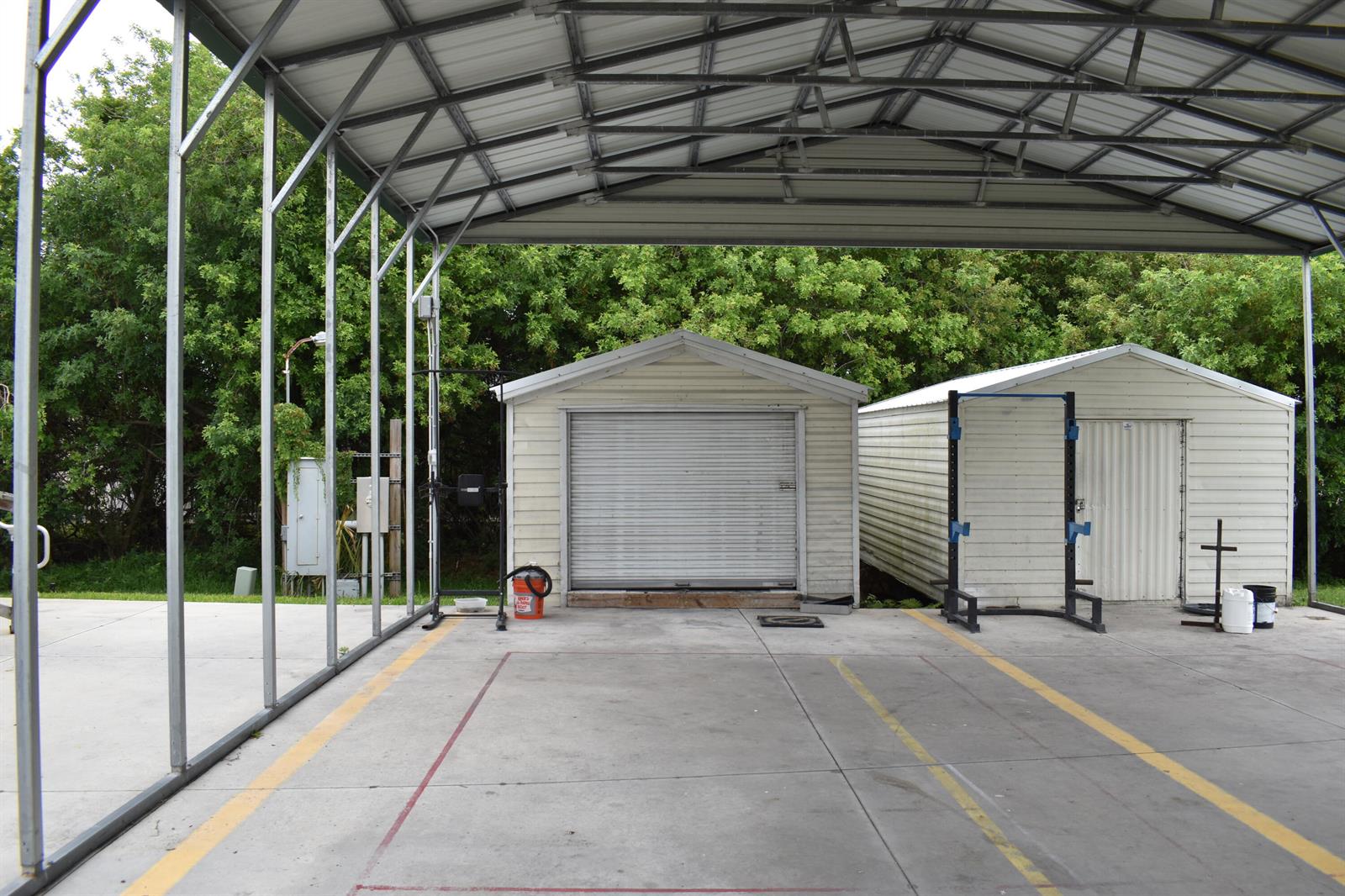 An open-air metal carport features two small storage sheds and two weightlifting racks on concrete — 1893 90th Avenue, Vero Beach FL 32966, VERO BEACH, FL