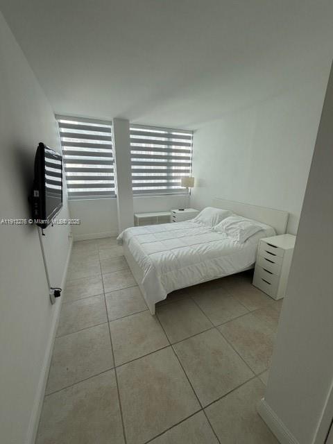 A bedroom with tile flooring, a white bed, a wall-mounted television, and striped window blinds — 5055 Collins Ave # 9A, Miami Beach FL 33140, MIAMI BEACH, FL