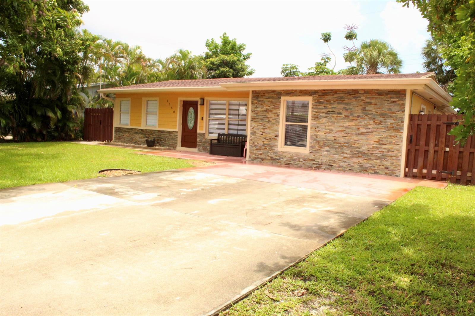 A single-story house features a stone facade, yellow siding, a concrete driveway, and wooden fences — 1558 NE 30th Street, Pompano Beach FL 33064, POMPANO BEACH, FL