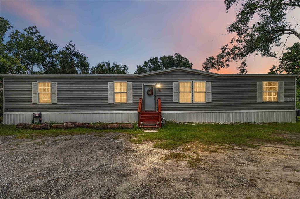A single-story manufactured home with grey siding, a red front porch, and a gravel driveway — 4235 PINE TREE DRIVE, ST CLOUD, FL