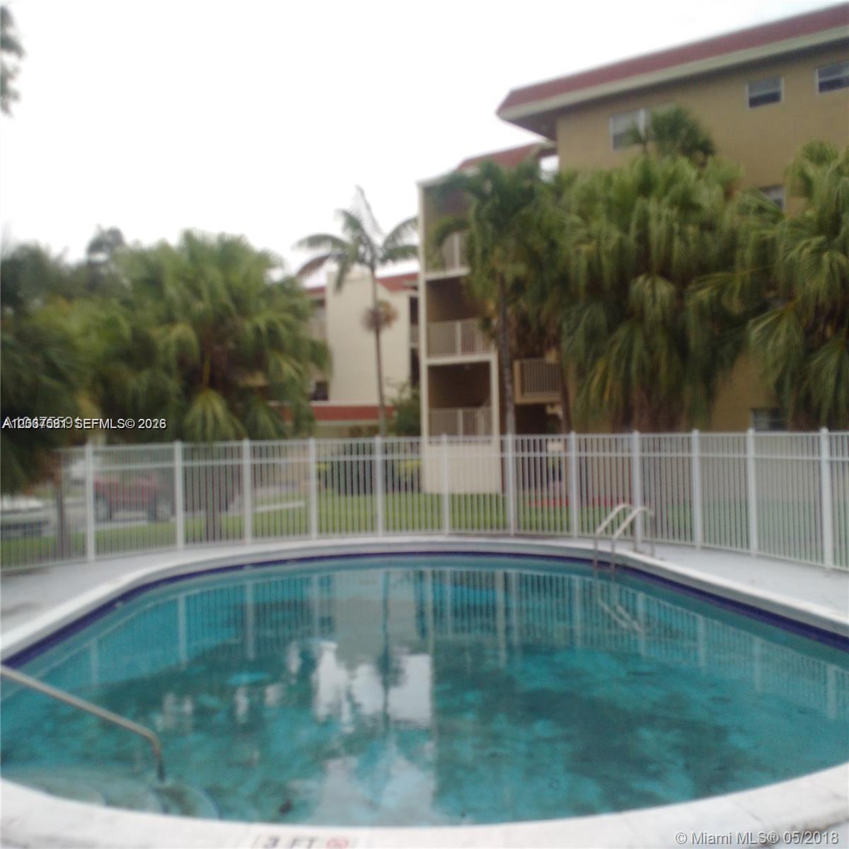 A swimming pool is enclosed by a white fence in front of a multi-story apartment building — SUNRISE, FL