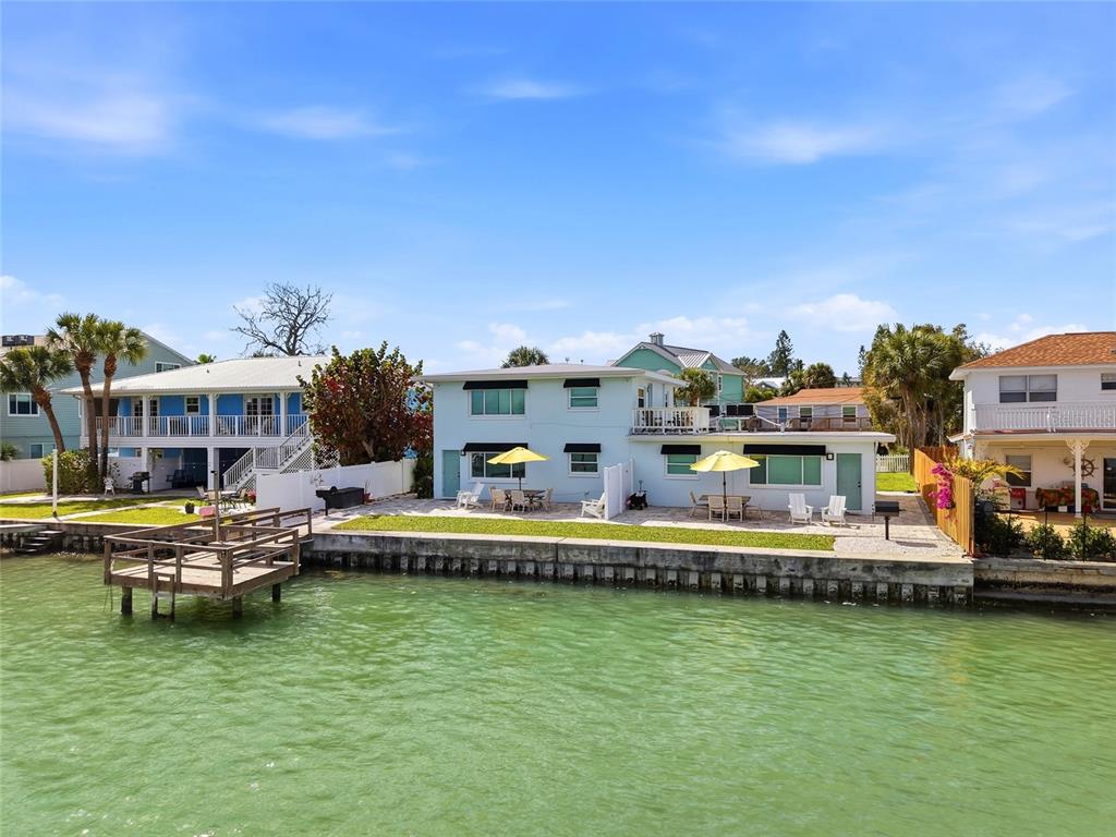 A light blue waterfront house features a patio, yellow umbrellas, and a wooden boat dock — 2605 BAY BOULEVARD, INDIAN ROCKS BEACH, FL