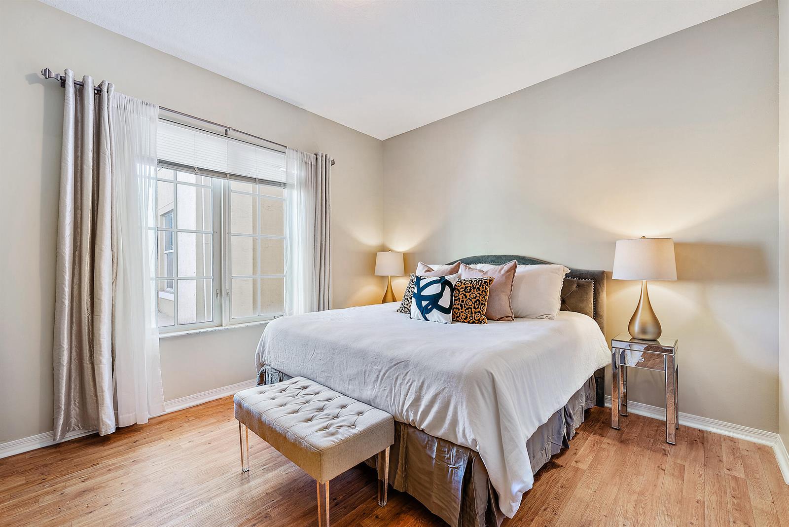 A bedroom features a king-sized bed, mirrored nightstands, a tufted bench, and wood-look flooring — 1200 Town Center Drive # 225, Jupiter FL 33458, JUPITER, FL