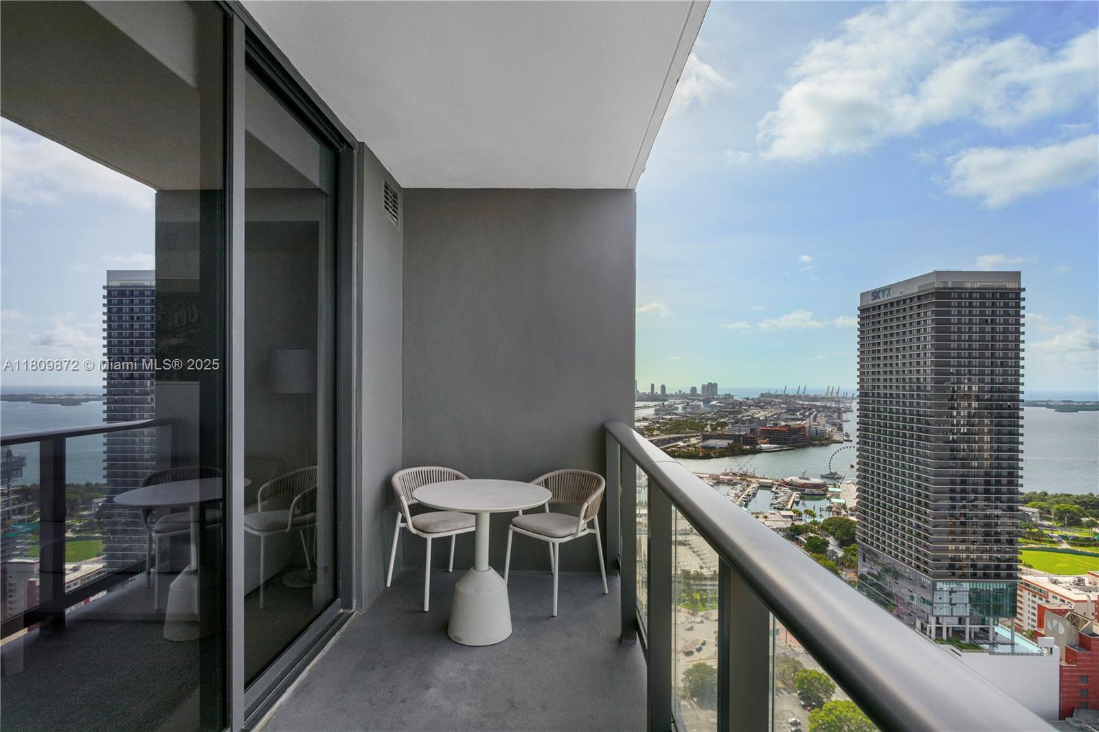A balcony with a small table and two chairs overlooks a city waterfront and skyscrapers — 601 NE 1st St # 3907, Miami FL 33132, MIAMI, FL