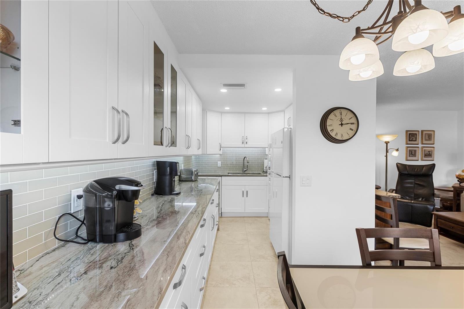 A white galley kitchen features granite countertops, light tile flooring, and a dining area nearby — 210 Oakridge N # 210, Deerfield Beach FL 33442, DEERFIELD BEACH, FL
