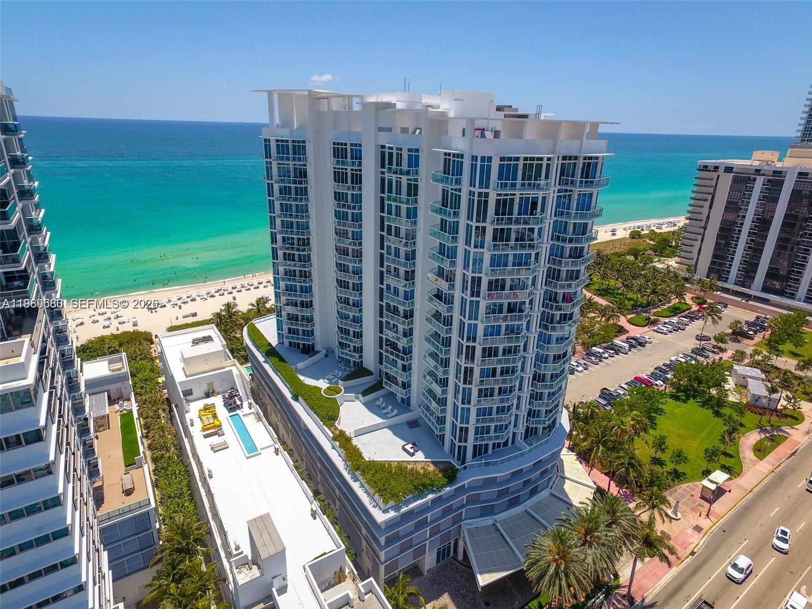 An aerial view shows a tall, modern white residential building situated next to a beach — 6515 Collins Ave # 1709, Miami Beach FL 33141, MIAMI BEACH, FL