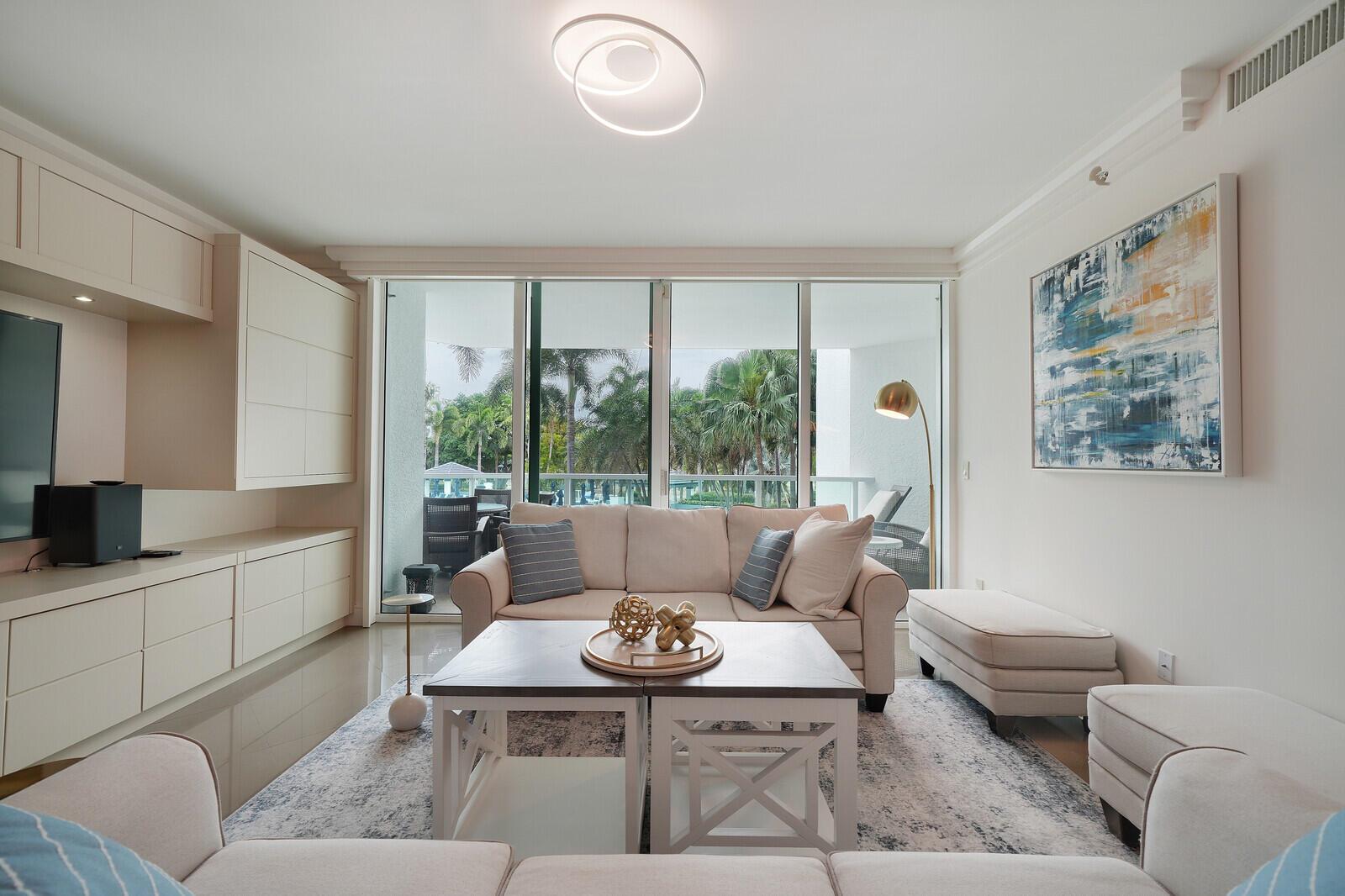 A bright living room features a beige sofa, coffee table, and large windows overlooking palm trees — 550 SE Mizner Boulevard # B209, Boca Raton FL 33432, BOCA RATON, FL