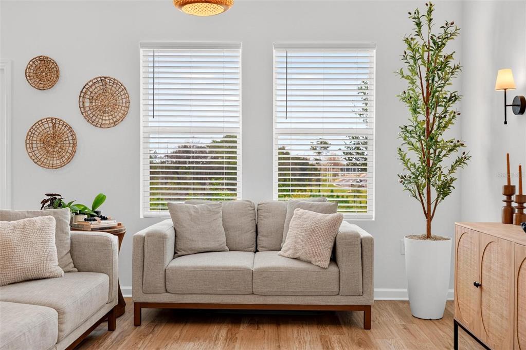 A living room features a light gray loveseat, two windows, a tall potted plant, and wall decor — 8704 SPORTSMAN LOOP, GROVELAND, FL