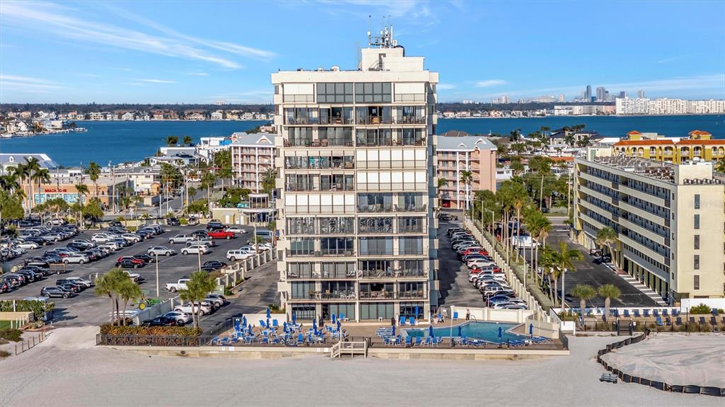 An aerial view shows a tall beachfront condominium building with a pool and parking lots — 5396 GULF BOULEVARD UNIT 407, ST PETE BEACH, FL