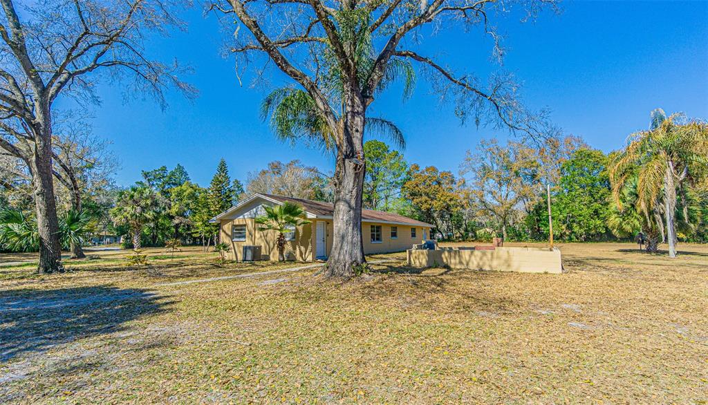 A single-story yellow house sits on a large, grassy lot with several mature trees — 3902 AUTUMN PALM DRIVE, ZEPHYRHILLS, FL