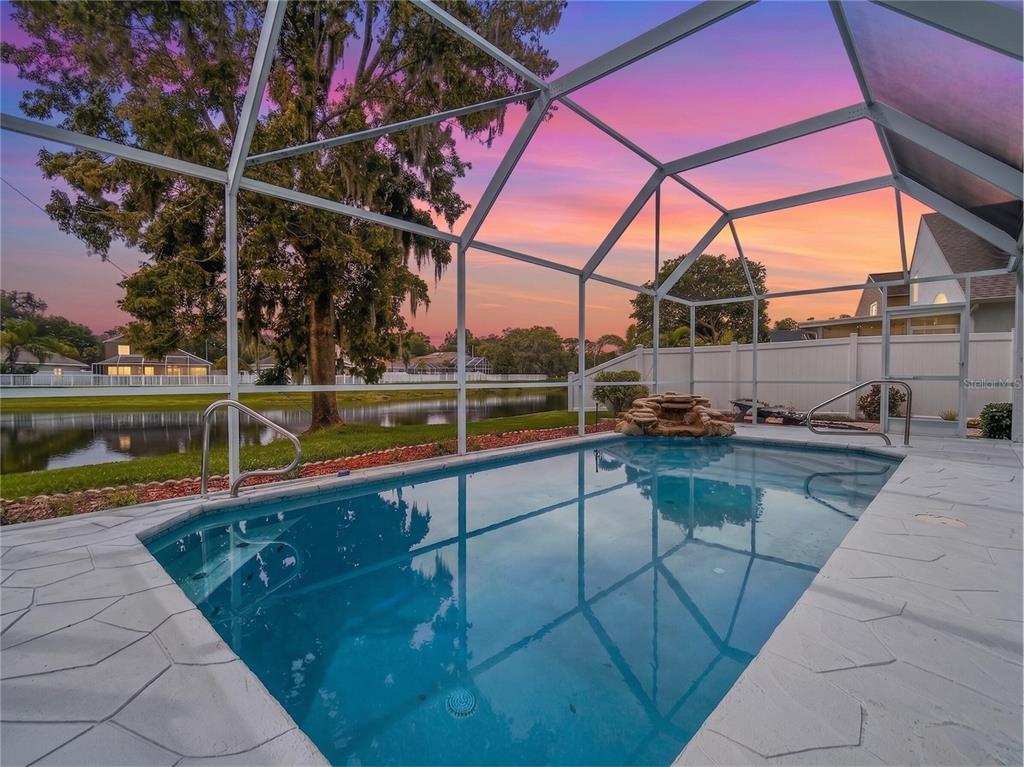 A screened-in swimming pool with a rock water feature overlooks a pond at sunset — 4842 WELLBROOK DRIVE, NEW PORT RICHEY, FL