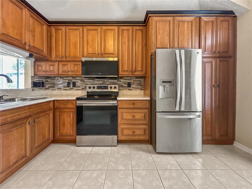A kitchen features wood cabinets, stainless steel appliances, a tiled backsplash, and light-colored floor tiles — 4842 WELLBROOK DRIVE, NEW PORT RICHEY, FL