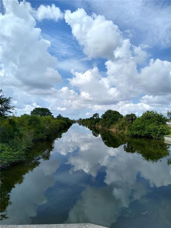 A canal flows between lush green vegetation under a bright blue sky with large clouds — 2710 NE 6TH AVENUE, CAPE CORAL, FL