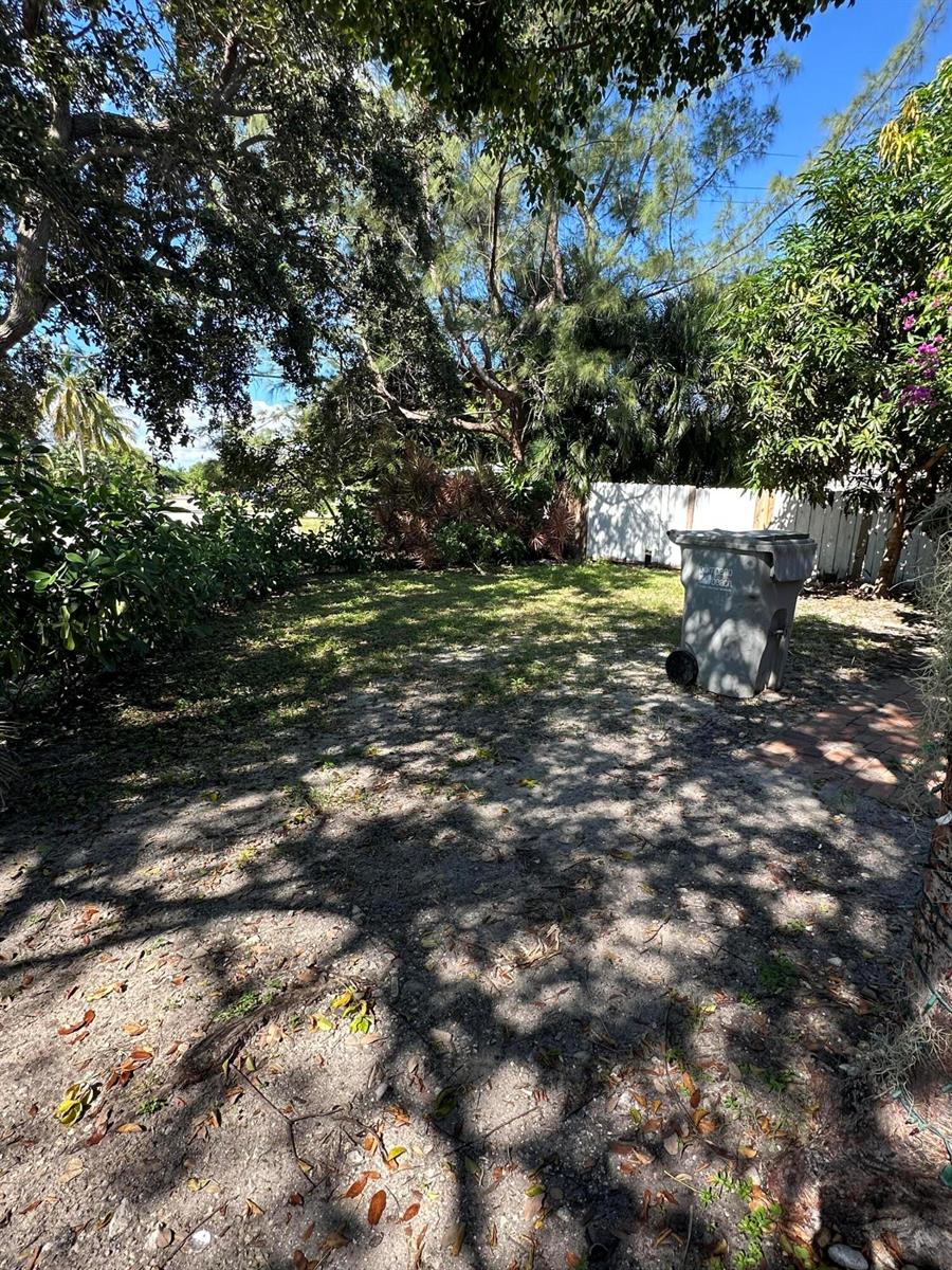 A backyard with a sandy lawn, mature trees, and a gray trash bin — 1571 NE 43rd Street, Pompano Beach FL 33064, POMPANO BEACH, FL
