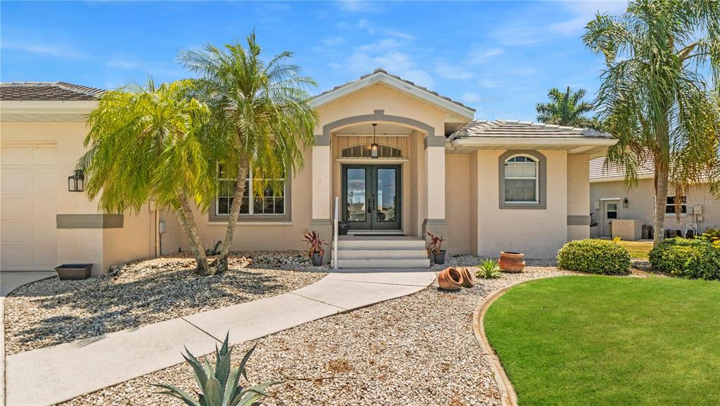 The exterior of a single-story beige house features a paved walkway, palm trees, and landscaping — 527 SAINT GIRONS COURT, PUNTA GORDA, FL