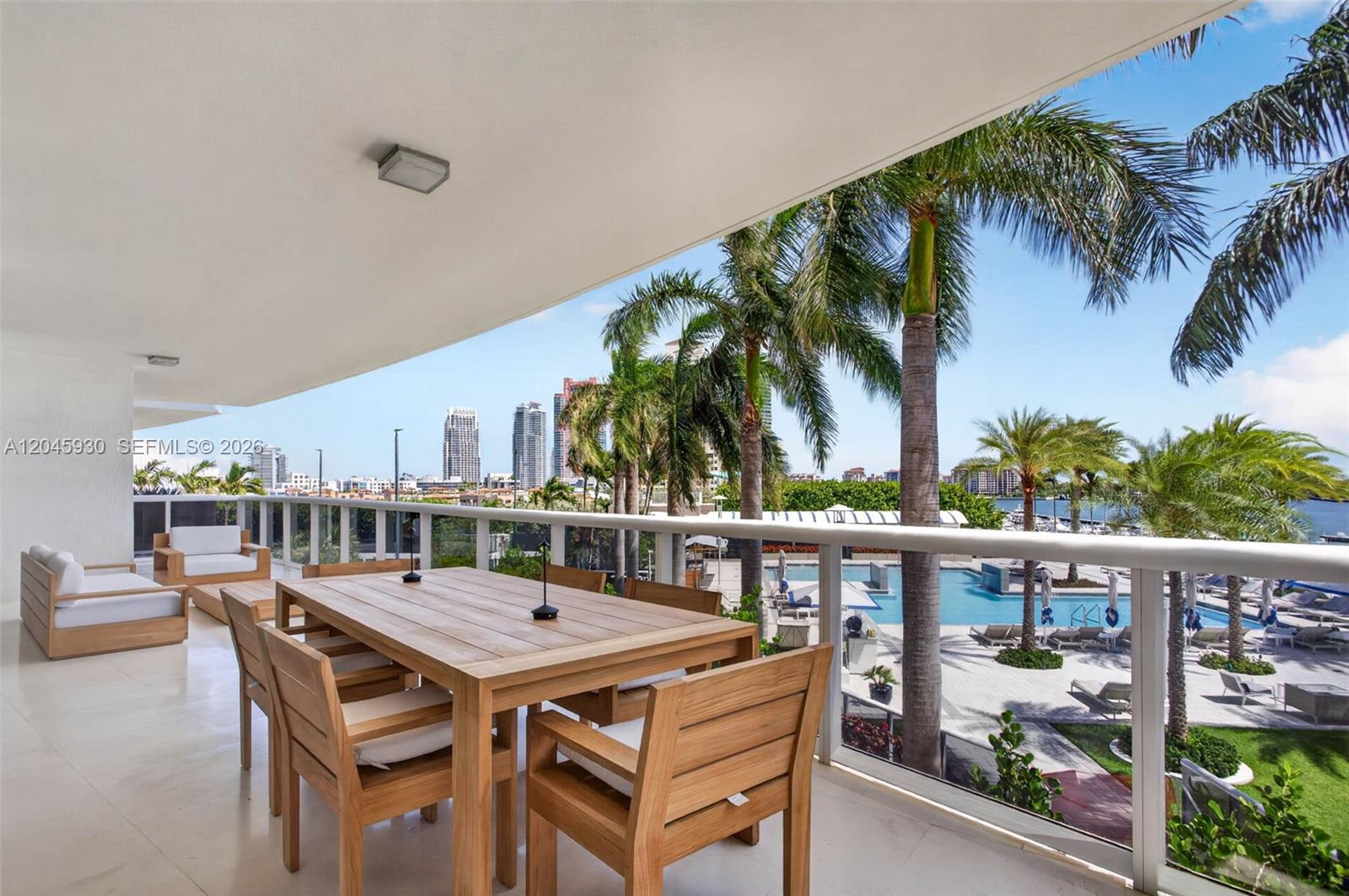 A balcony features a wooden dining set and lounge chairs overlooking a pool and city — 400 Alton Rd # 706, Miami Beach FL 33139, MIAMI BEACH, FL