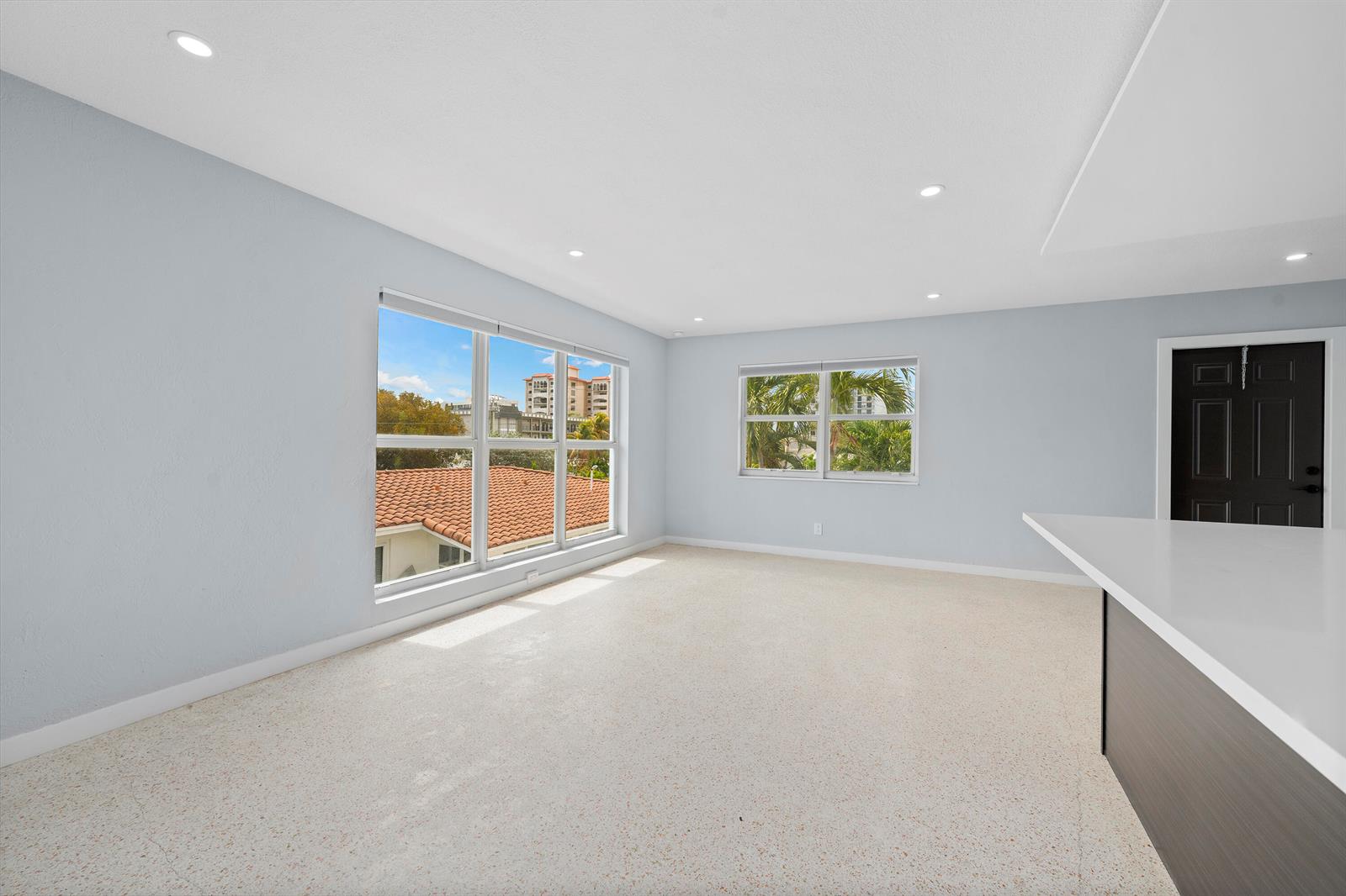 An empty room with light blue walls, terrazzo flooring, large windows, and a black door — 3220 NE 7th Place # 3, Pompano Beach FL 33062, POMPANO BEACH, FL