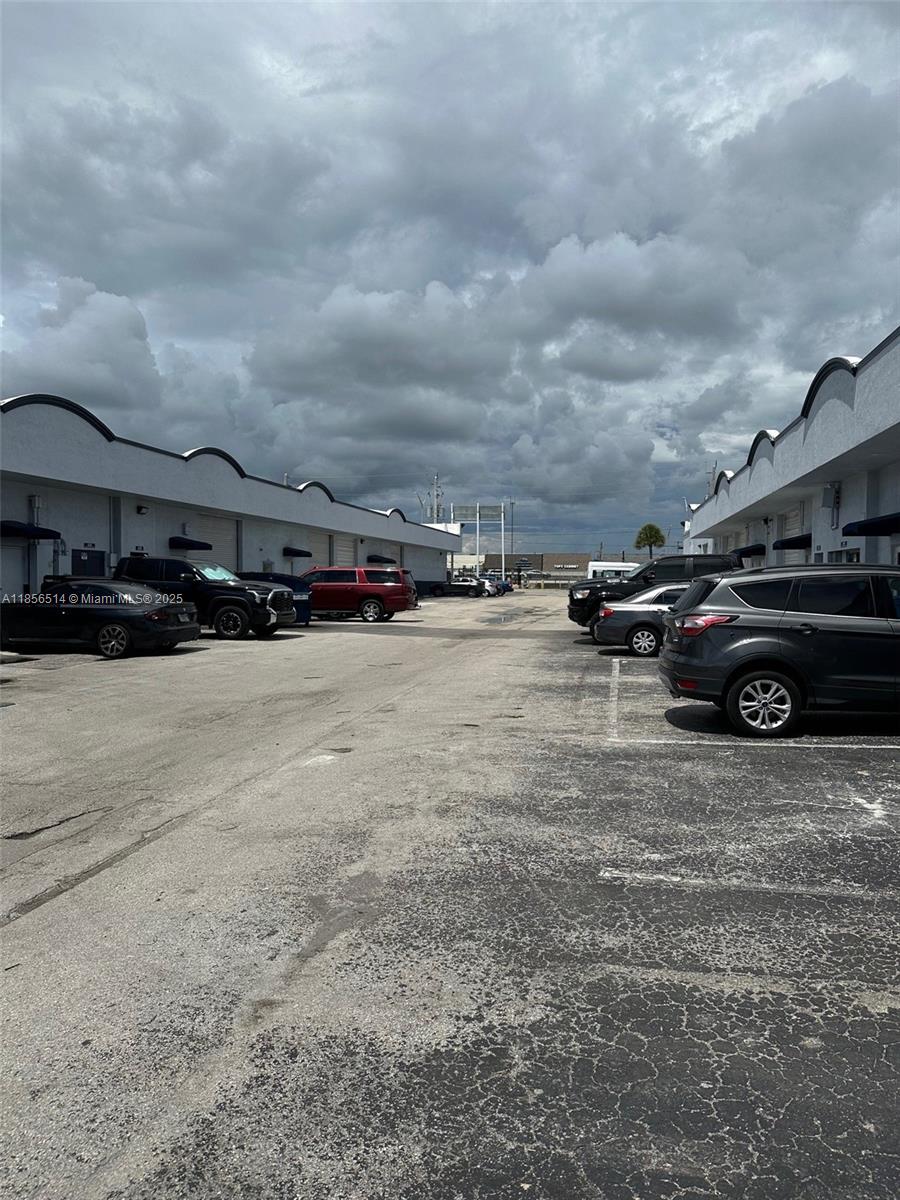 An exterior view of a commercial parking lot between two rows of industrial warehouse buildings — 470 Ansin Blvd # 470J, Hallandale Beach FL 33009, HALLANDALE BEACH, FL