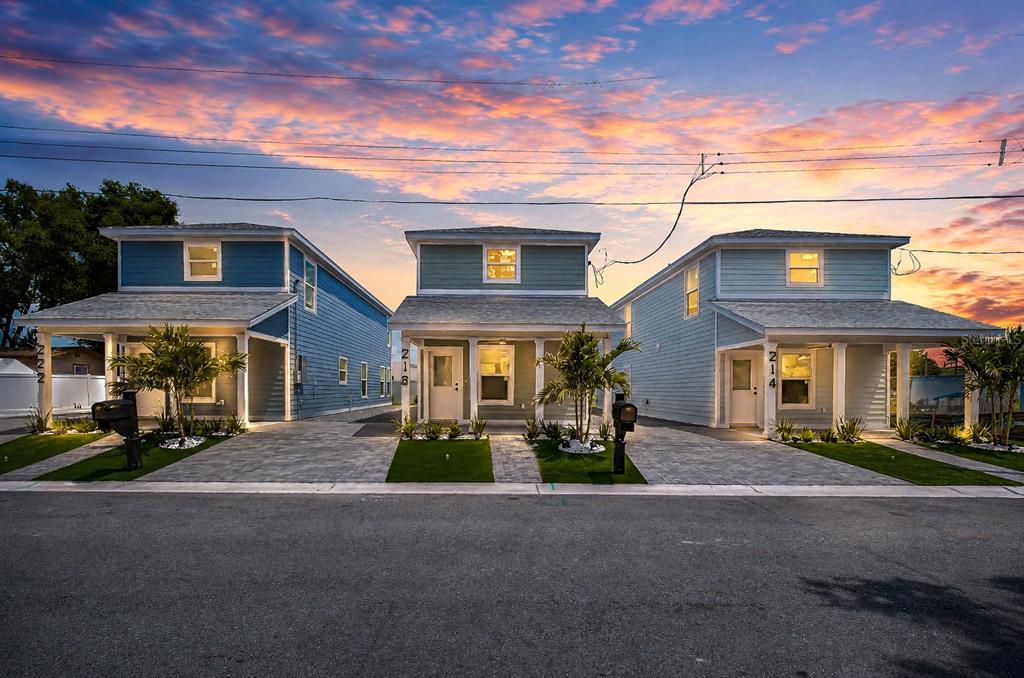 Three identical two-story houses with blue siding and paved driveways stand under a sunset sky — 218 8TH AVENUE DRIVE W, BRADENTON, FL