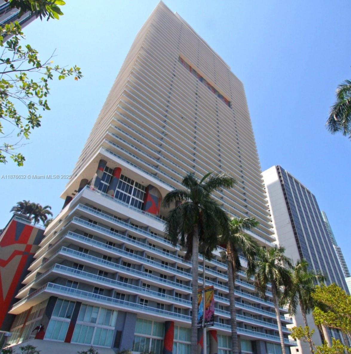 A low-angle view of a tall, modern high-rise building with balconies and palm trees — 50 Biscayne Blvd # 711, Miami FL 33132, MIAMI, FL