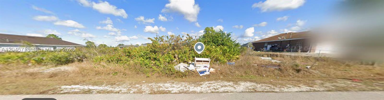 A vacant, overgrown residential lot with scattered debris sits between two houses under a blue sky — 2806 54TH ST W, Lehigh Acres FL 33971, LEHIGH ACRES, FL