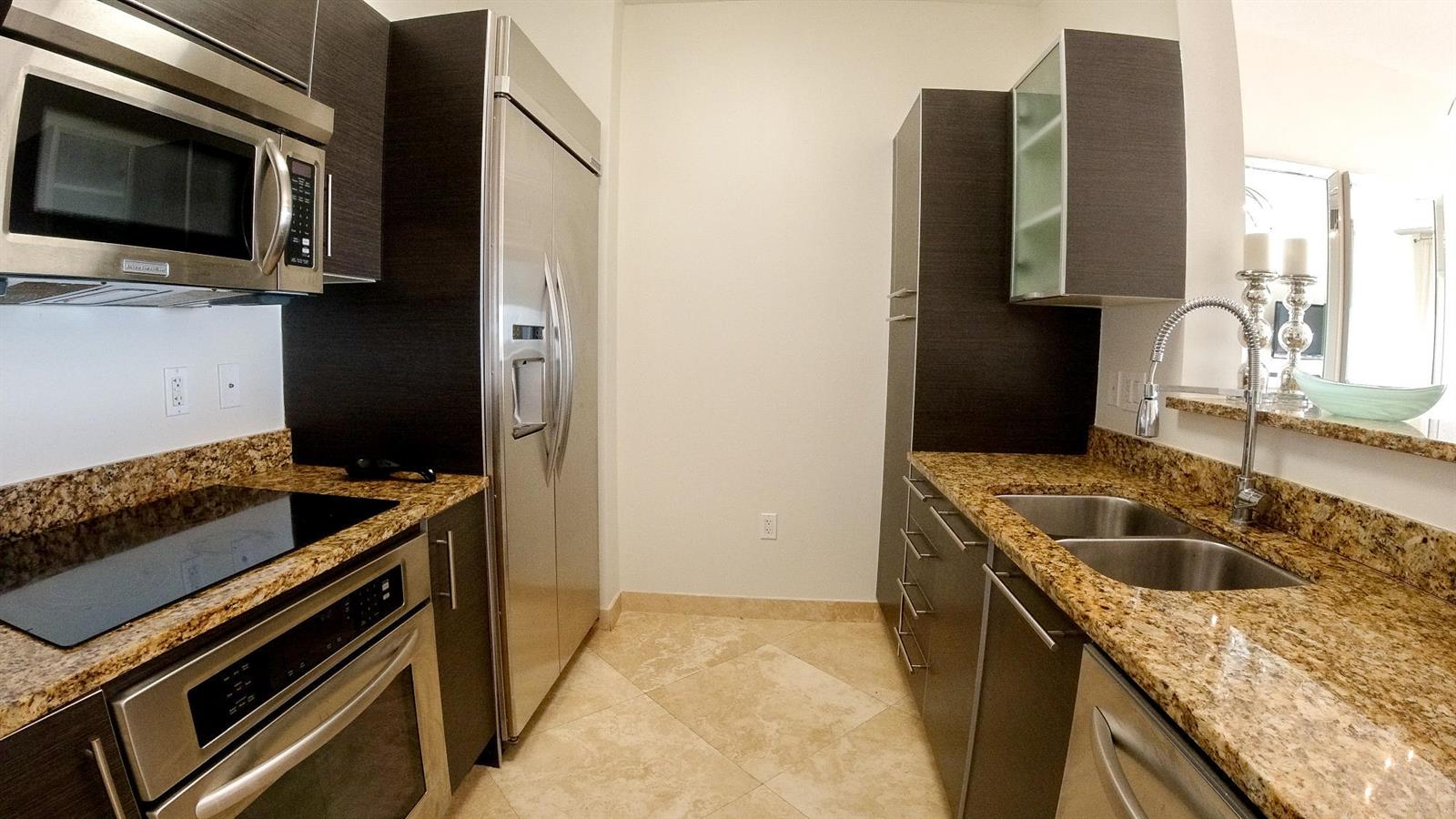 A galley kitchen features dark wood cabinets, granite countertops, and stainless steel appliances — 550 Okeechobee Boulevard # 1721, West Palm Beach FL 33401, WEST PALM BEACH, FL
