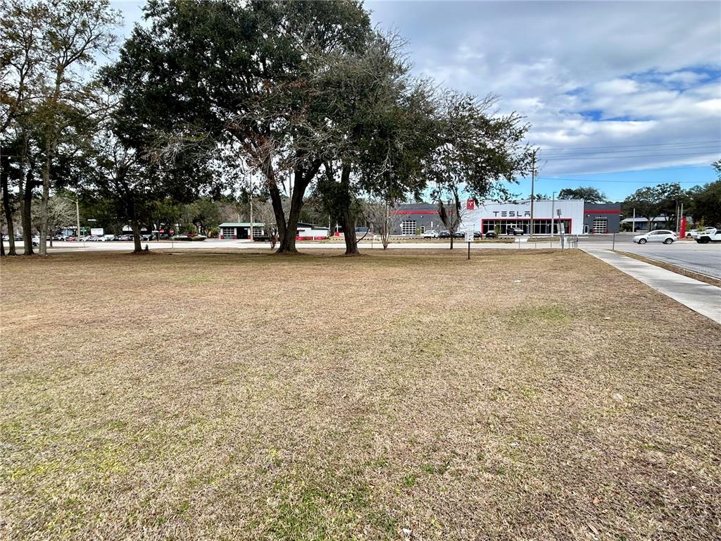 A vacant, grassy lot sits in front of a Tesla service center and mature trees — 2550 N MAIN STREET, GAINESVILLE, FL