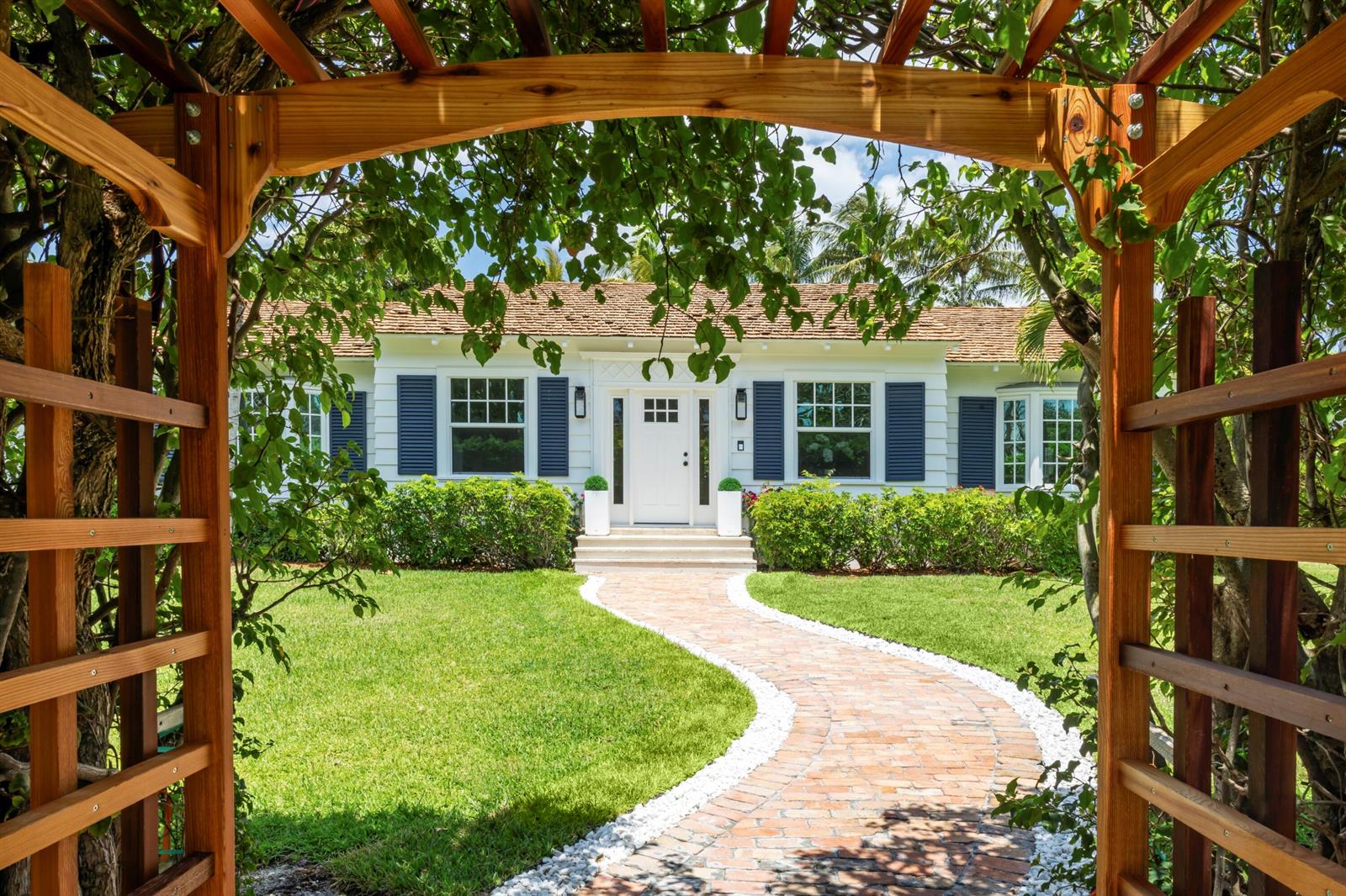 A white house with blue shutters is viewed through a wooden garden arbor and walkway — 1017 Tamarind Road, Delray Beach FL 33483, DELRAY BEACH, FL