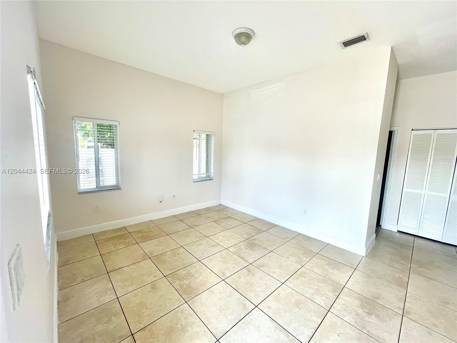 An empty room features beige tile flooring, white walls, two windows, and white louvered closet doors — 2487 W 5th Ct, Hialeah FL 33010, HIALEAH, FL