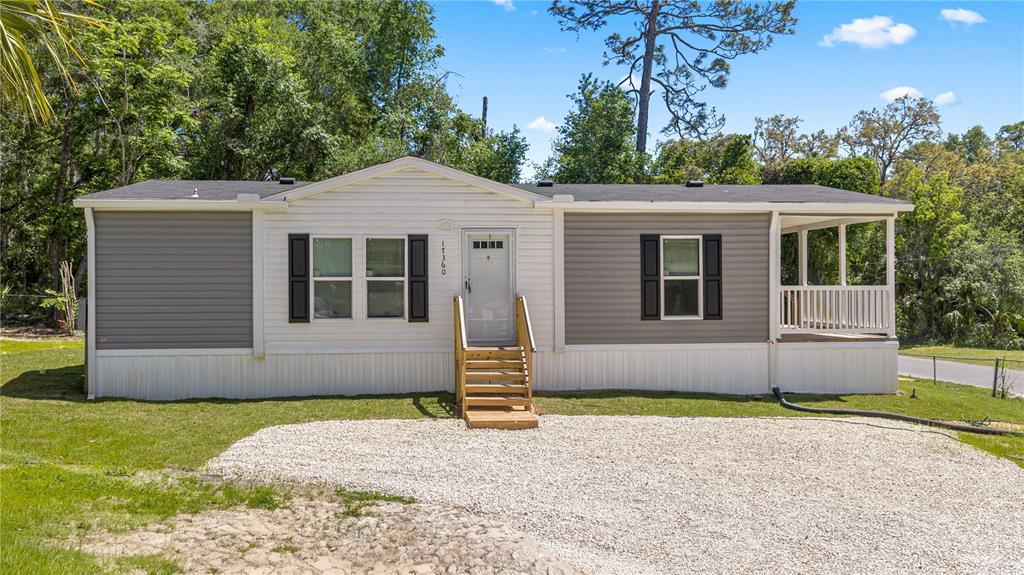 A single-story manufactured home features gray and white siding, a front porch, and gravel driveway — 17360 SE 65TH STREET, OCKLAWAHA, FL